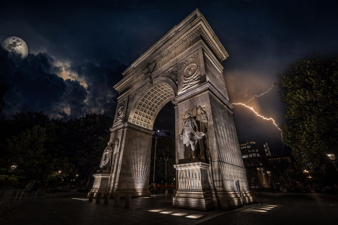 Historic stone triumphal arch in a city park at night, lit by spotlights highlighting carved statues and reliefs, with dramatic storm clouds, a full moon and a lightning bolt in the sky.
