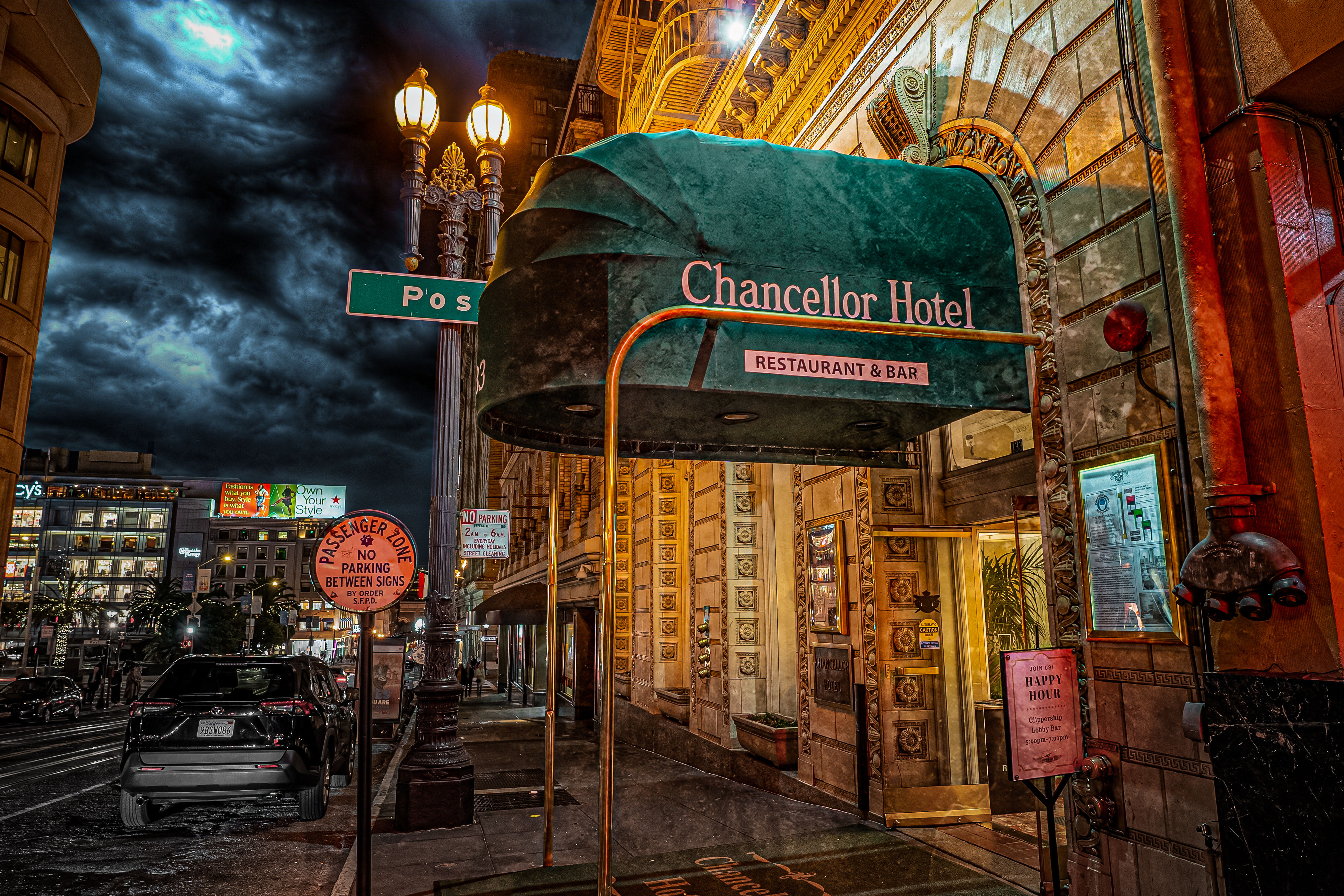 Downtown hotel entrance at night with a green canopy labeled 'Restaurant & Bar', ornate golden facade and vintage street lamps under a dramatic stormy sky, parked cars and city street scene.