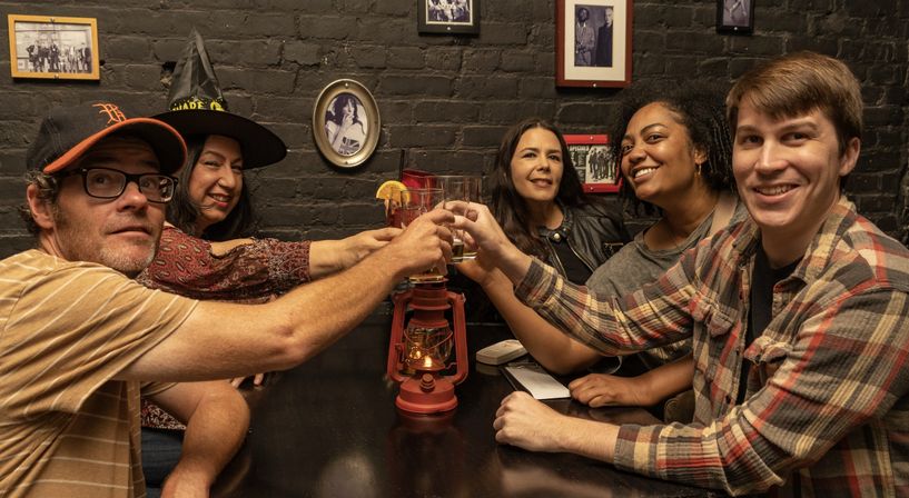 Five friends smiling and toasting with drinks at a cozy bar table, exposed black brick wall with framed photos, red lantern centerpiece and one person wearing a witch hat.