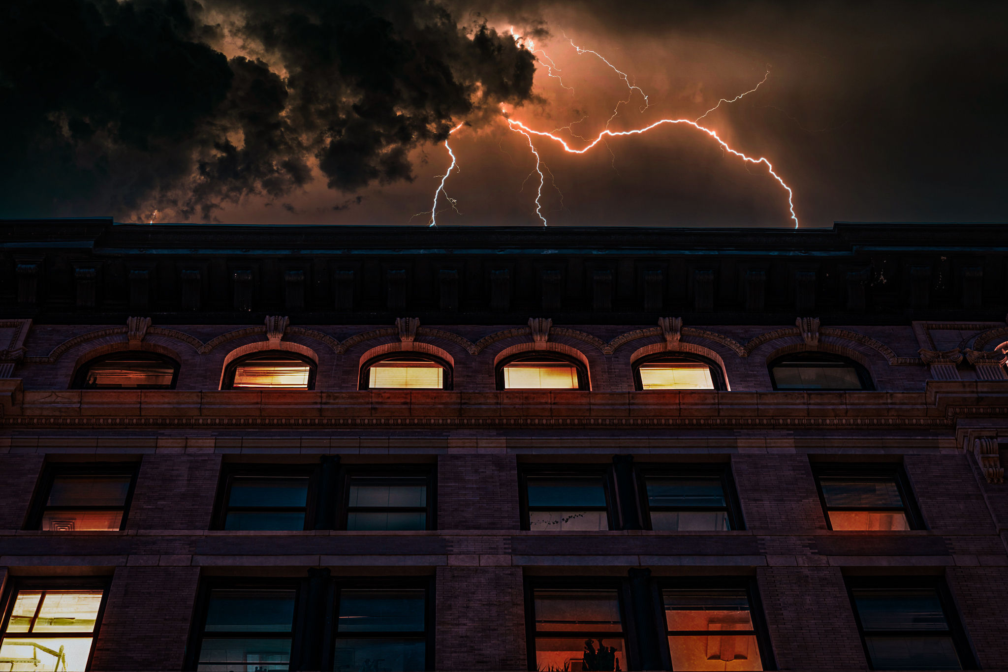 Lightning bolts streak across a stormy night sky above a historic brick downtown building with glowing windows — dramatic urban thunderstorm scene