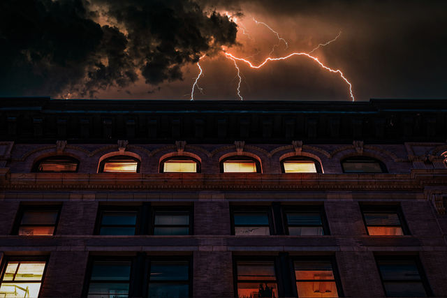 Lightning bolts streak across a stormy night sky above a historic brick downtown building with glowing windows — dramatic urban thunderstorm scene