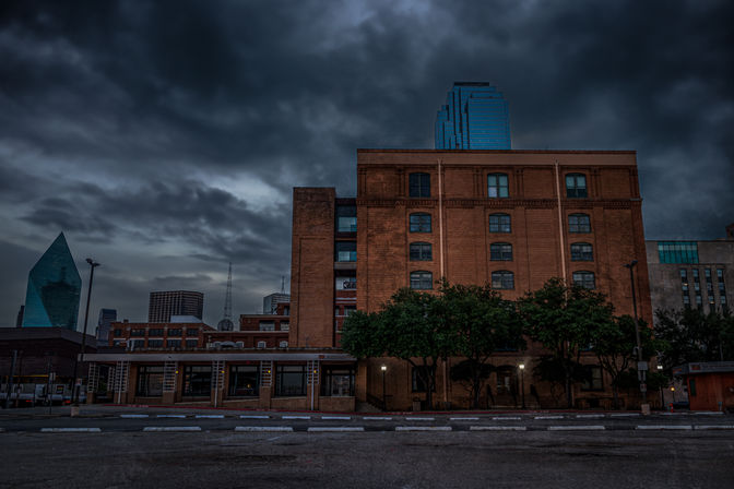 Moody urban scene at dusk: historic red-brick warehouse and trees in front of modern blue-glass skyscrapers under dramatic storm clouds.