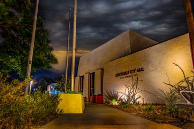 Night view of a modern Southwestern city hall building with illuminated stucco facade, tall flagpoles, desert plants and a dramatic cloudy sky.