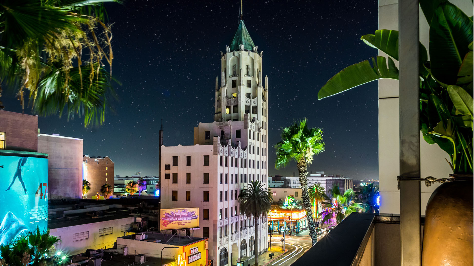 Rooftop view of a palm-lined Los Angeles street at night with a glowing Art Deco tower, vibrant neon storefronts, and a starry sky.