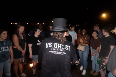 Tour guide in a top hat and black shirt leading an outdoor nighttime ghost tour, holding a lantern while a diverse group of smiling young adults listens on a dark street.