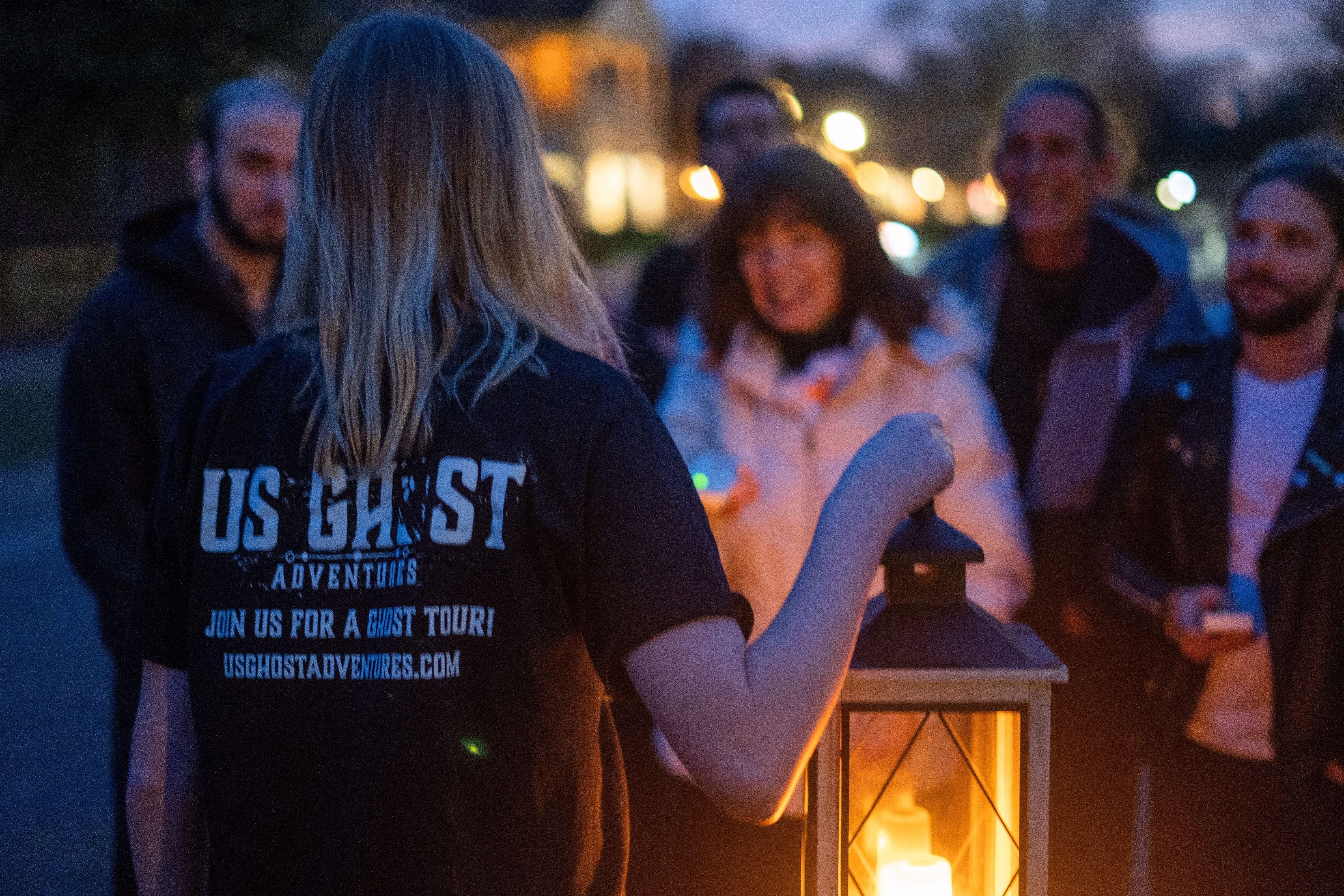 Lantern-bearing tour guide leading a small, smiling group on an evening ghost tour down a dimly lit street at dusk.