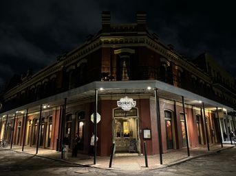 Night view of a historic red-brick corner restaurant with wrought-iron balcony and covered sidewalk, warmly lit on a quiet French Quarter street in New Orleans.