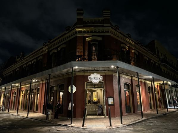 Nighttime street view of a historic red-brick corner restaurant with wrought-iron balcony in New Orleans' French Quarter, warm interior lights and a few people on the sidewalk.