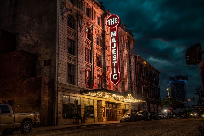 Historic downtown theater with tall vertical neon marquee and glowing canopy, ornate stone façade and parked cars on a moody nighttime city street