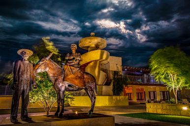 Bronze sculptures of a standing man and a woman riding a mule in a lit Southwestern plaza at night, with a spiral outdoor staircase and dramatic stormy clouds overhead.