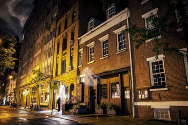 Nighttime downtown street with historic brick rowhouses and storefronts, warm yellow streetlights glowing on wet pavement and a pedestrian-crossing sign in view.