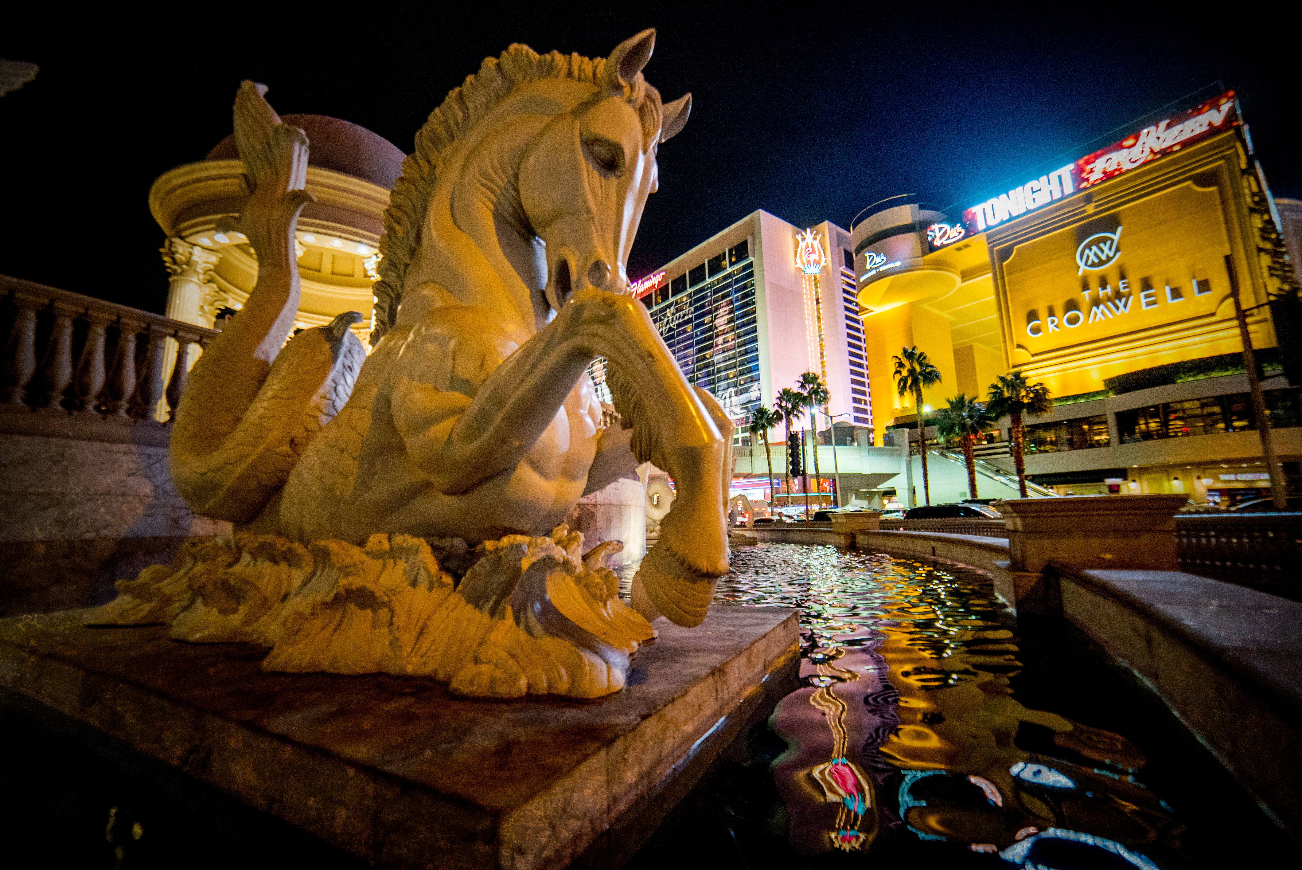 Stone mythical seahorse fountain beside a reflecting pool on the Las Vegas Strip at night, neon-lit hotels and palm trees reflected in the water.