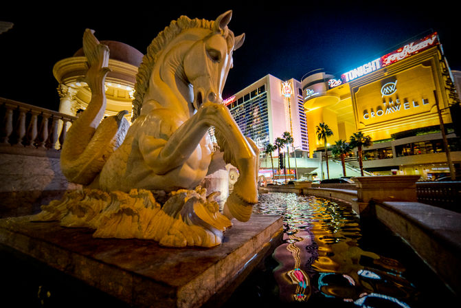 Stone mythical seahorse fountain beside a reflecting pool on the Las Vegas Strip at night, neon-lit hotels and palm trees reflected in the water.