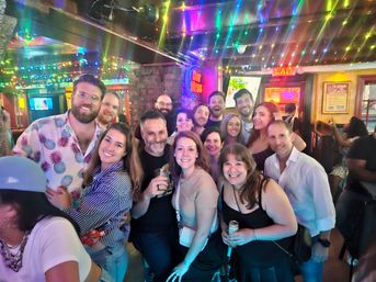 Smiling group of friends posing at a lively neighborhood bar with colorful string lights, neon signs, exposed brick, and drinks