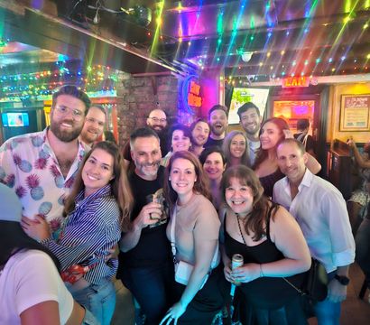 Large group of friends smiling for a selfie inside a lively bar at night, colorful string lights and neon signs overhead, people holding drinks and mingling.