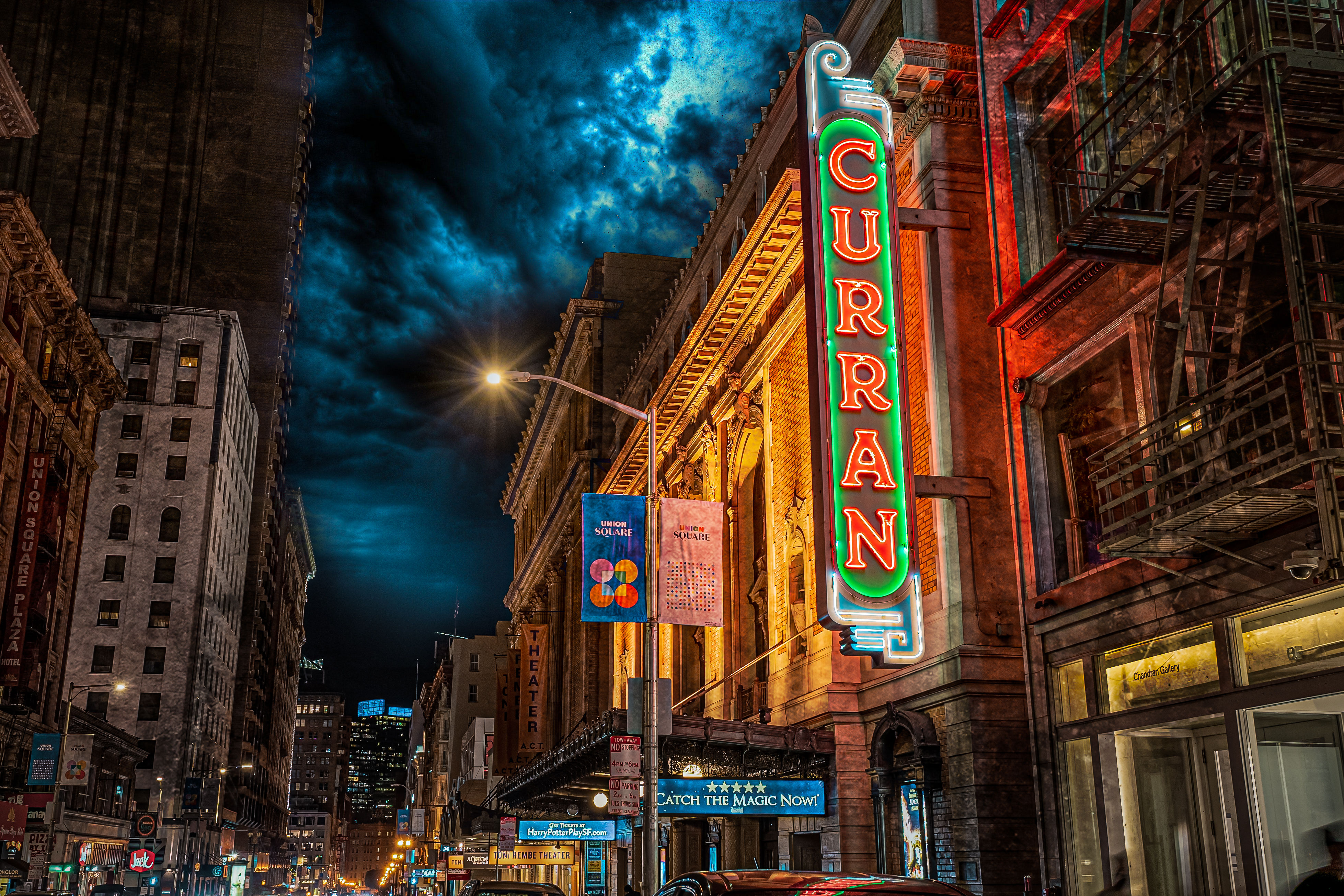 Vibrant downtown nighttime street with a historic theater facade and tall vertical neon marquee glowing above cars, lit street lamps and banners under a dramatic cloud-filled sky.