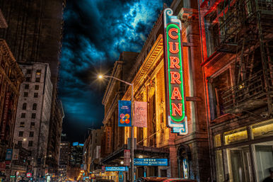 Vibrant downtown nighttime street with a historic theater facade and tall vertical neon marquee glowing above cars, lit street lamps and banners under a dramatic cloud-filled sky.