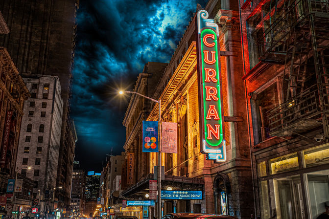 Vibrant downtown nighttime street with a historic theater facade and tall vertical neon marquee glowing above cars, lit street lamps and banners under a dramatic cloud-filled sky.