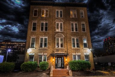 Four-story historic brick apartment building at night with warm-lit entrance, trimmed shrubs, and a dramatic stormy sky above downtown high-rises.