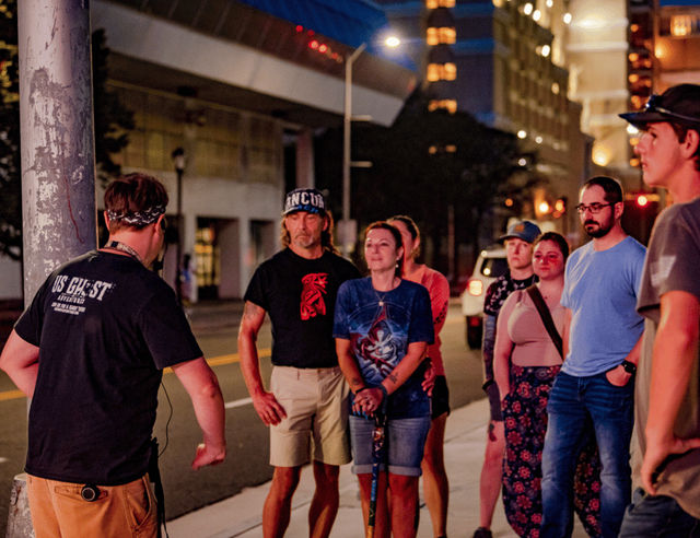 Group of adults gathered on a downtown sidewalk at night listening to a guide with a headset, with glowing city lights and tall buildings in the background.