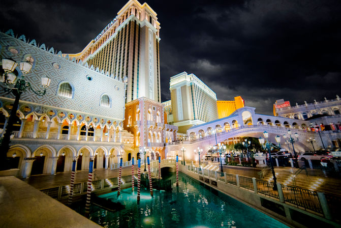 Nighttime Las Vegas Strip scene of a Venetian-style hotel facade with arched colonnades, illuminated bridge over a turquoise canal with striped gondola poles under a dramatic cloudy sky.