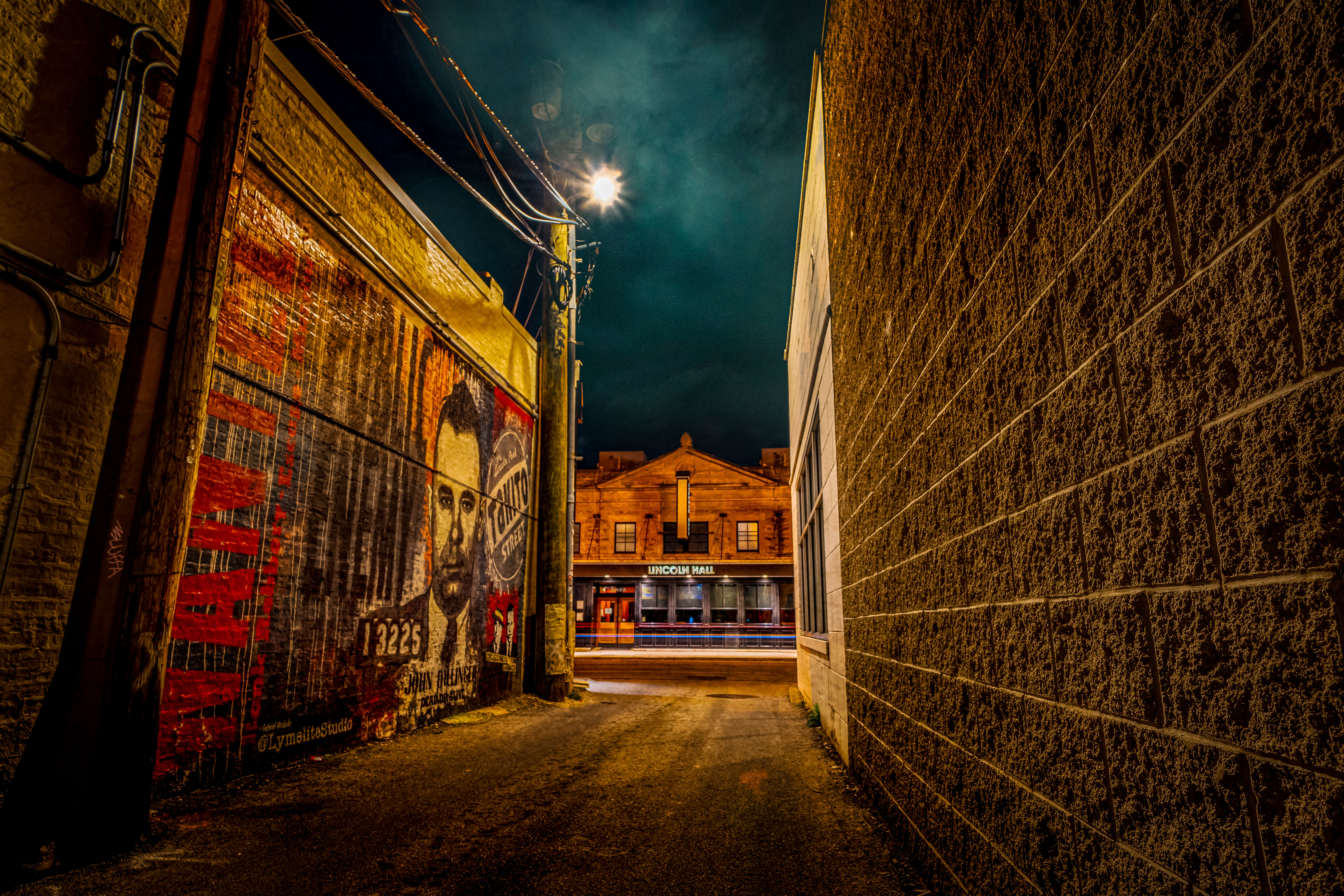 Moody nighttime urban alley with textured brick walls, a colorful mural portrait on the left, a glowing streetlight overhead, and a warmly lit historic-style building across the street.