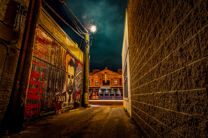 Moody nighttime urban alley with textured brick walls, a colorful mural portrait on the left, a glowing streetlight overhead, and a warmly lit historic-style building across the street.