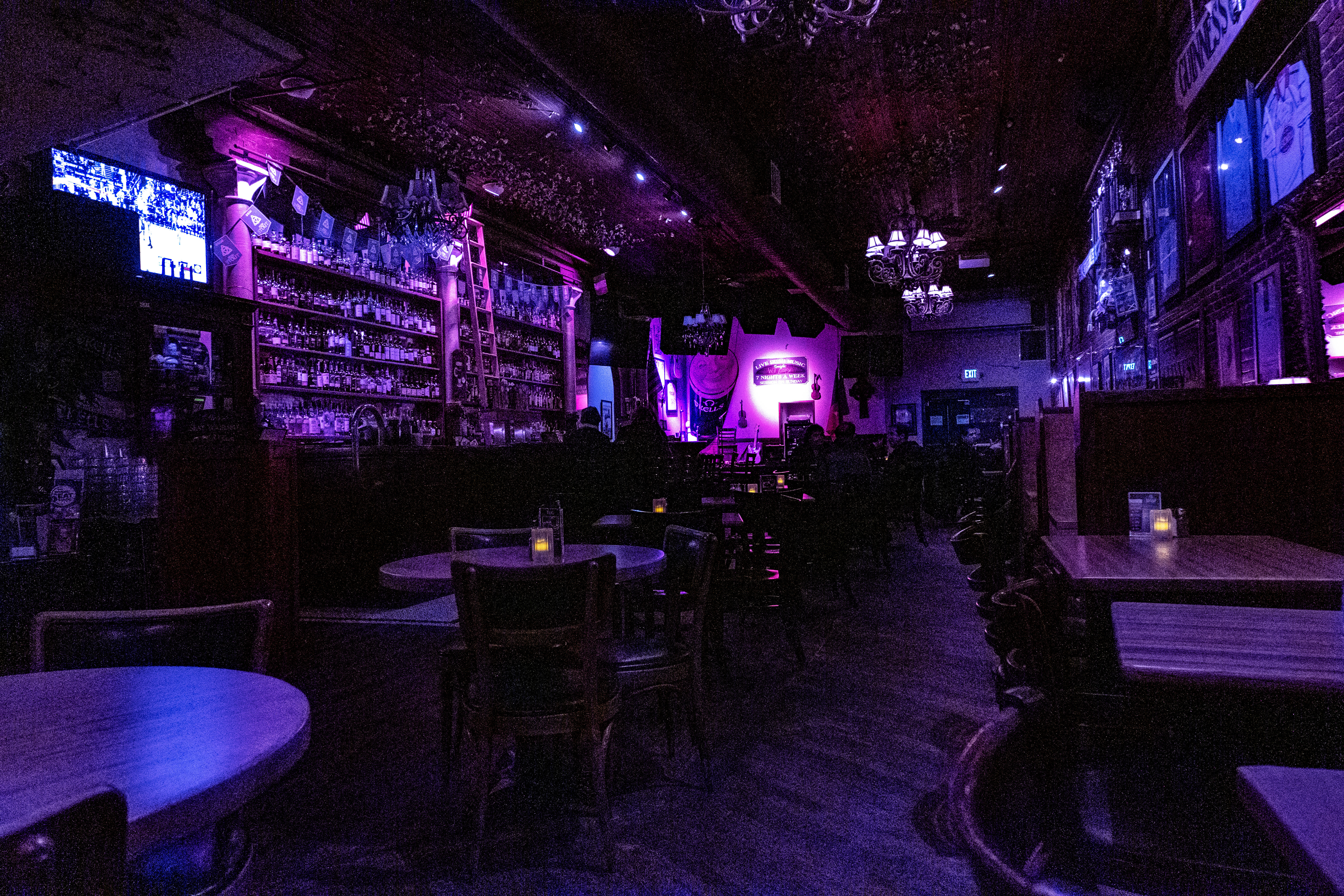 Dimly lit live music bar interior bathed in purple neon, empty wooden tables and chairs with candle-lit votives, long back bar stocked with bottles and a ladder, chandeliers overhead and a small stage with instruments in the background — moody nightlife scene.