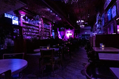 Dimly lit live music bar interior bathed in purple neon, empty wooden tables and chairs with candle-lit votives, long back bar stocked with bottles and a ladder, chandeliers overhead and a small stage with instruments in the background — moody nightlife scene.