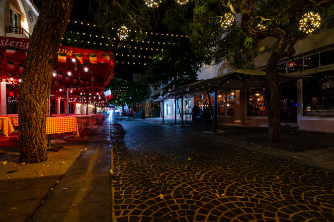 Nighttime downtown pedestrian street with cobblestone-patterned pavement, outdoor dining under red awnings and checkered tablecloths, trees and hanging string lights creating a cozy, festive urban atmosphere.
