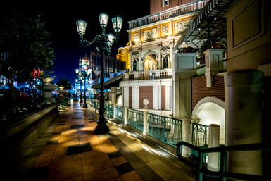 Nighttime urban promenade with Venetian-style stone facades and arches, vintage lamp posts casting warm pools of light on a patterned tile walkway and decorative green railings.