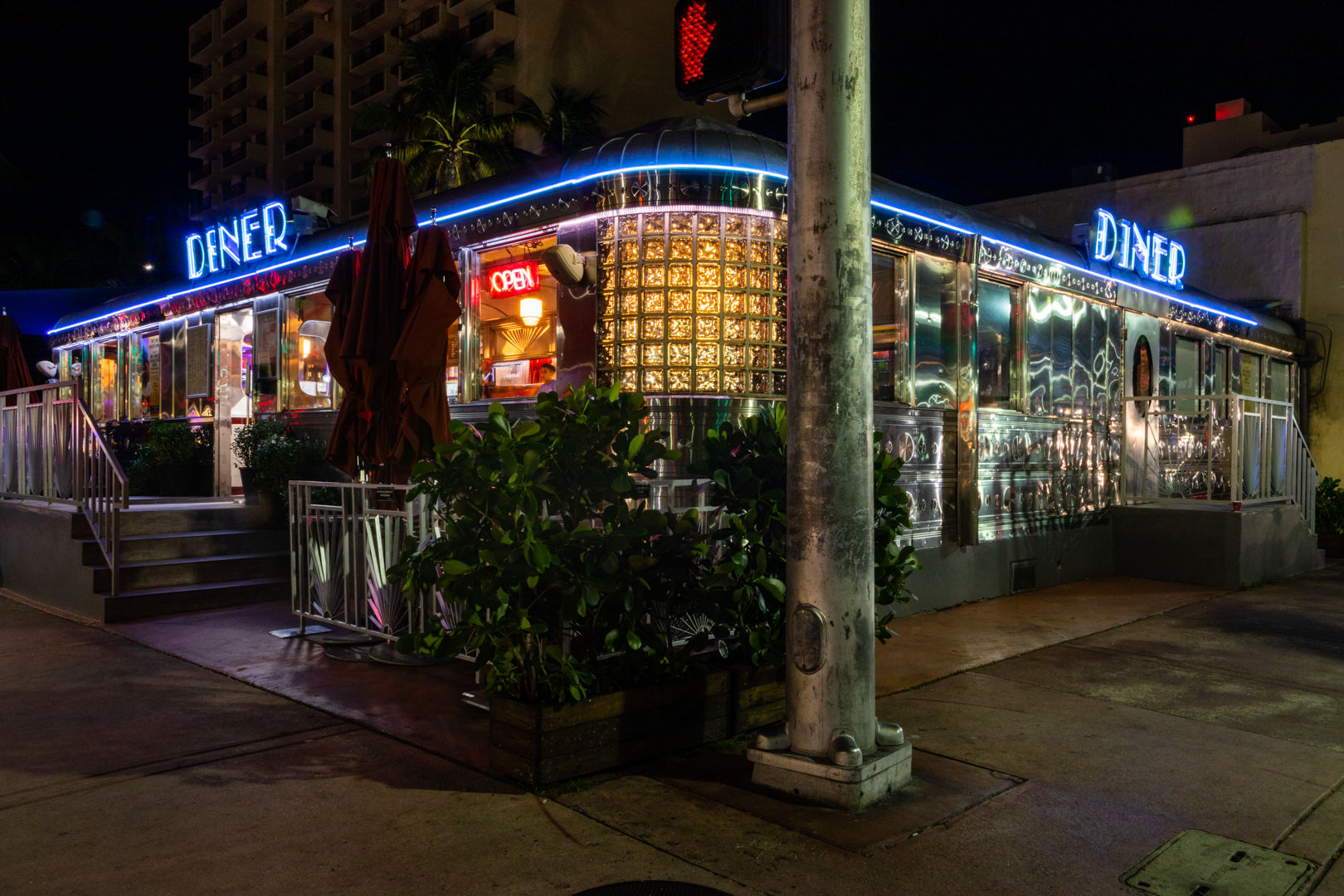 Retro stainless-steel diner at night with blue neon 'DINER' sign and glowing 'OPEN' window, outdoor umbrellas and plants on an urban sidewalk