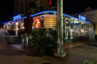 Retro stainless-steel diner at night with blue neon 'DINER' sign and glowing 'OPEN' window, outdoor umbrellas and plants on an urban sidewalk