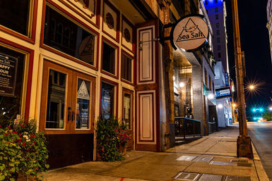 Cozy, warmly lit restaurant entrance on a quiet downtown city street at night with a wood-paneled facade, round hanging sign, planted flower boxes and glowing streetlights along the sidewalk.