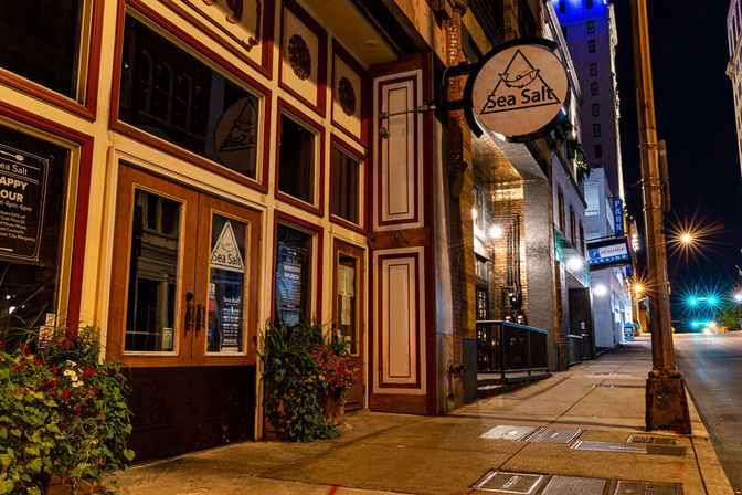 Cozy, warmly lit restaurant entrance on a quiet downtown city street at night with a wood-paneled facade, round hanging sign, planted flower boxes and glowing streetlights along the sidewalk.