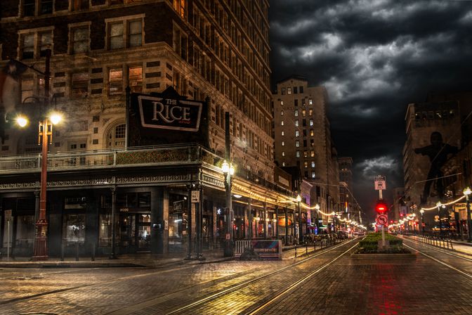 Moody nighttime scene in a historic downtown: wet brick street and streetcar tracks flanked by lit storefronts and vintage lampposts under dramatic clouds.