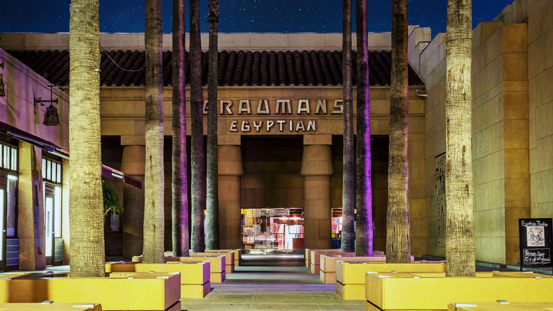 Night courtyard of an Egyptian‑style theater — tall palm trees, purple uplighting, yellow planter benches and a glowing central entrance.