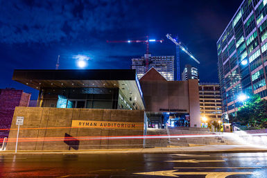 Night view of a downtown Nashville concert hall entrance under a moonlit sky, framed by construction cranes, glass high-rises and wet street light trails.