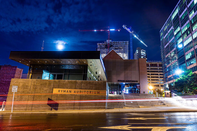 Night view of a downtown Nashville concert hall entrance under a moonlit sky, framed by construction cranes, glass high-rises and wet street light trails.