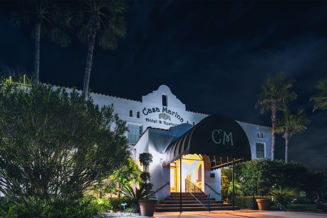 Nighttime view of a Mediterranean‑style white hotel entrance with an illuminated arched canopy, inviting warm glow, stone steps, potted plants and palm trees under a dark sky.