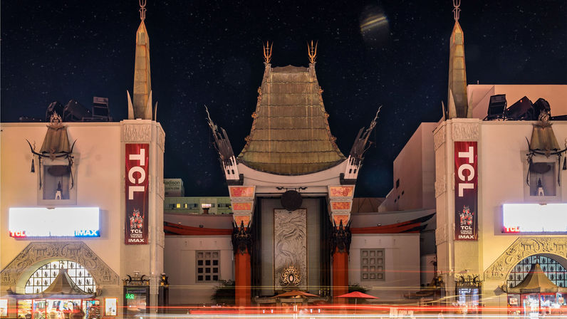 Night shot of an ornate Chinese-style movie palace façade on Hollywood Boulevard, Los Angeles — green pagoda roof, carved pillars, illuminated marquees and streaking car light trails