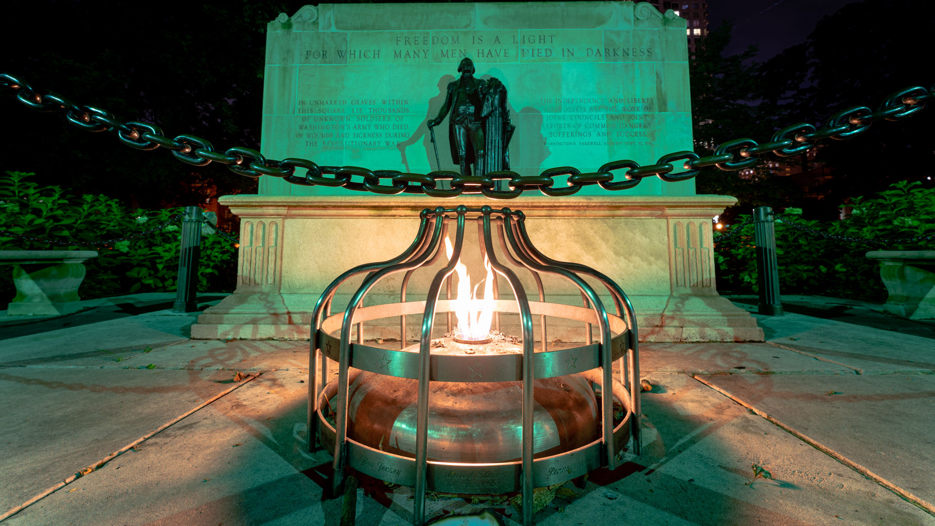 Night shot of a glowing eternal flame in a metal cage at the base of a stone memorial with a bronze statue and heavy chain, green-tinted park lighting.