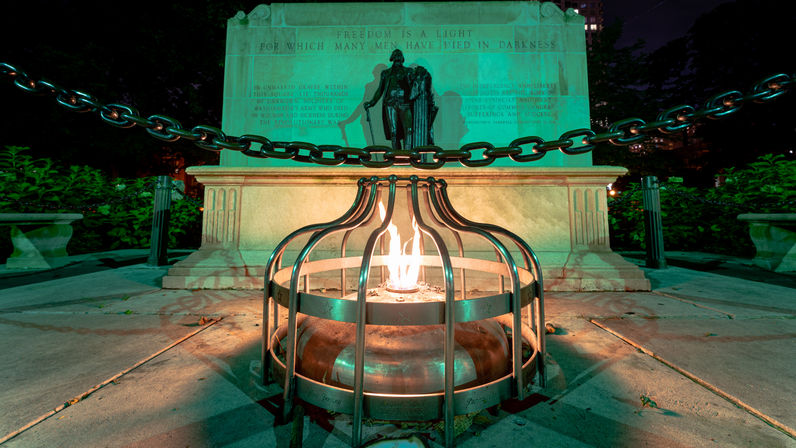 Night shot of a glowing eternal flame in a metal cage at the base of a stone memorial with a bronze statue and heavy chain, green-tinted park lighting.