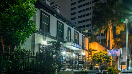 Nighttime urban hotel exterior with glowing neon "VIP" sign, two-story white facade, outdoor terrace with railings and chairs, palm trees and high-rise buildings in the background