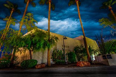 Nighttime view of a low‑slung stucco public library surrounded by desert landscaping and tall palm trees, lit by warm exterior lights beneath dramatic stormy blue clouds
