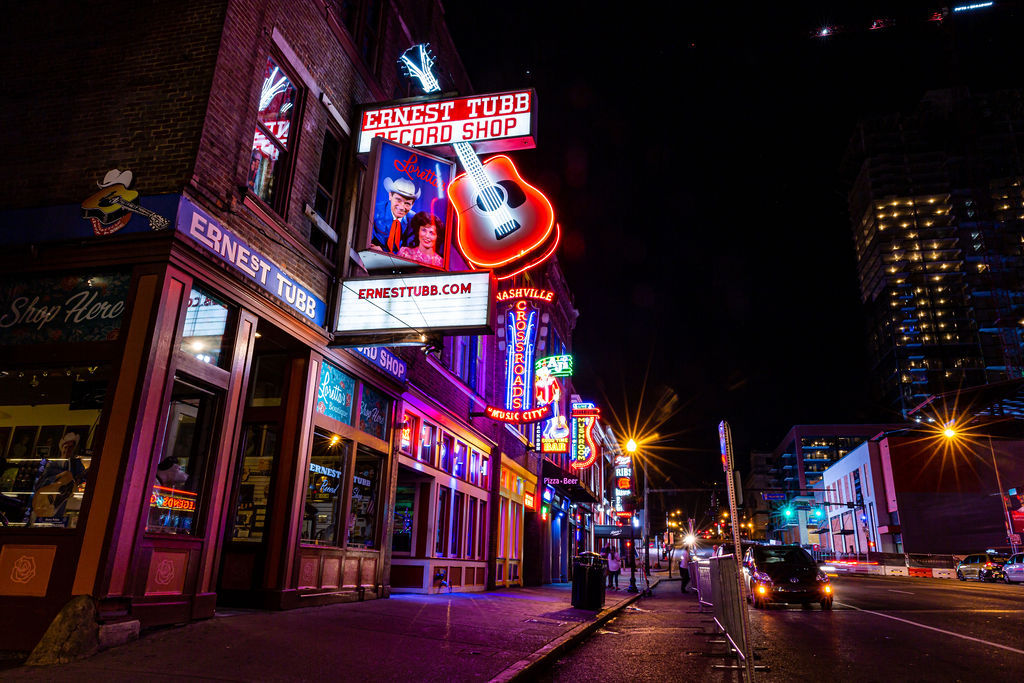 Vibrant neon-lit downtown street at night featuring a music-themed record shop with a giant glowing guitar sign, colorful storefronts, and bustling nightlife.