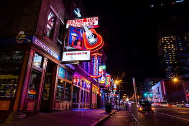 Vibrant neon-lit downtown street at night featuring a music-themed record shop with a giant glowing guitar sign, colorful storefronts, and bustling nightlife.