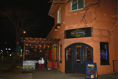 Nighttime city corner brick restaurant-bar with a covered outdoor patio strung with warm string lights, sidewalk signs advertising brunch and happy hour, and an illuminated arched entrance.