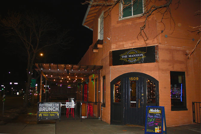 Nighttime city corner brick restaurant-bar with a covered outdoor patio strung with warm string lights, sidewalk signs advertising brunch and happy hour, and an illuminated arched entrance.