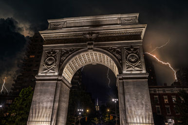 Illuminated Washington Square Arch in New York City at night, dramatic storm clouds and lightning bolts framing the stone arch with a distant lit skyscraper on the skyline.